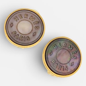 May include: A pair of round gold-tone earrings featuring a dark, iridescent mother-of-pearl inlay. Each earring is engraved with the text HERMÈS PARIS in a circular pattern, accented by small diamond-shaped symbols on the left and right sides.