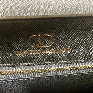 Puede incluir: Primer plano de un bolso de mano de cuero negro con cremallera dorada y el logotipo dorado de Valentino Garavani. El cuero tiene una apariencia texturizada.