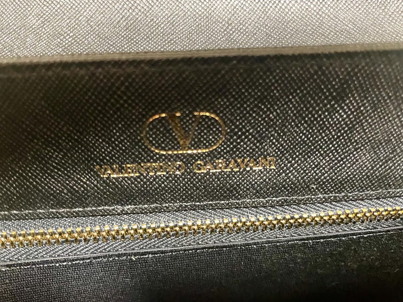 Puede incluir: Primer plano de un bolso de mano de cuero negro con cremallera dorada y el logotipo dorado de Valentino Garavani. El cuero tiene una apariencia texturizada.