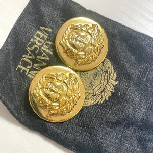 May include: A pair of gold-tone oval-shaped earrings with a raised Medusa head design. The earrings are in a black velvet pouch with the word "VERSACE" printed on it.