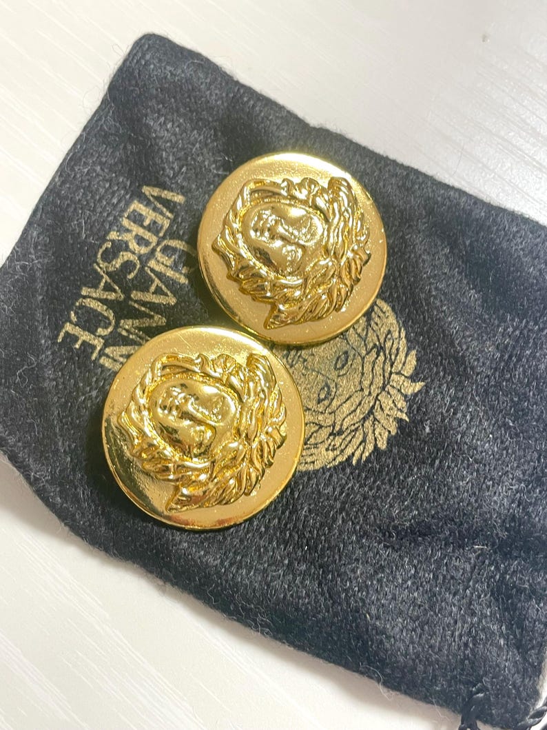 May include: A pair of gold-tone oval-shaped earrings with a raised Medusa head design. The earrings are in a black velvet pouch with the word "VERSACE" printed on it.
