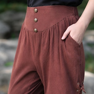 Op de afbeelding: Bruine broek met hoge taille, brede tailleband met drie decoratieve metalen knopen, een zijzak en een klein veterdetail aan de onderkant. De stof heeft een textuur en de tailleband is voorzien van een elastisch gedeelte.