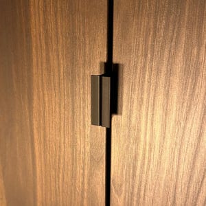 Könnte beinhalten: Ein mattschwarzer Schrankgriff aus Metall, der vertikal zwischen zwei dunkelbraunen Schranktüren mit Holzmaserung montiert ist. Der Griff hat ein rechteckiges Profil mit einer mittigen Nut und bildet ein funktionales Beschlagelement.