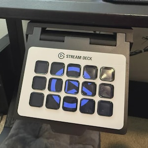 Könnte beinhalten: Ein schwarzer Stream Deck Controller, der unter einer Tischplatte montiert ist. Das Gerät verfügt über eine weiße Frontplatte mit fünfzehn quadratischen Tasten in einem Raster, die blaue Kreissymbole und ein Play-Symbol auf den Tasten zeigen.