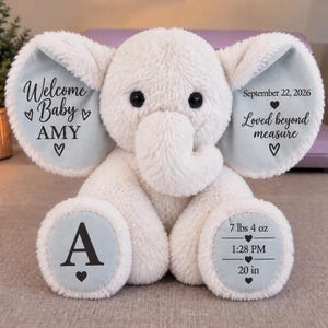 May include: A cream-coloured plush elephant toy with light blue ear and foot accents. The left ear reads Welcome Baby AMY, the right ear reads September 22, 2026 Loved beyond measure, the left foot has the letter A, and the right foot lists 7 lbs 4 oz, 1:28 PM, and 20 in.