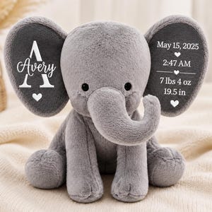 May include: A soft grey plush elephant toy with embroidered white text on its ears. The left ear features the name Avery with a large letter A and a heart. The right ear displays May 15, 2025, 2:47 AM, 7 lbs 4 oz, and 19.5 in with hearts.