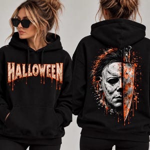 May include: A black hooded sweatshirt featuring the word HALLOWEEN in a distressed orange font on the front. The back displays a graphic illustration of a masked face partially obscured by a large knife with orange paint splatter effects.