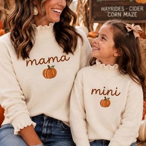 Könnte beinhalten: Zwei cremefarbene Langarmpullover mit gerüschten Stehkragen und Ärmelbündchen. Der größere Pullover zeigt den Schriftzug "mama" über einer Kürbisgrafik, während der kleinere Pullover den Schriftzug "mini" über einem passenden Kürbis zeigt.