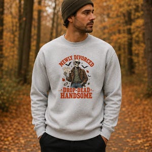 May include: A light gray crewneck sweatshirt featuring a graphic of a skeleton wearing sunglasses and a brown jacket, holding a book. The text reads "NEWLY DIVORCED" and "DROP-DEAD HANDSOME" in orange, with small bats and autumn leaves surrounding the figure.