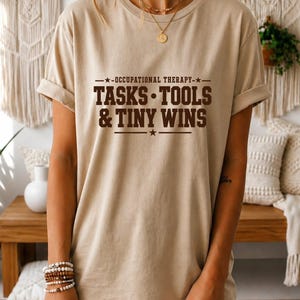 May include: A beige short-sleeve t-shirt featuring dark brown text that reads OCCUPATIONAL THERAPY TASKS TOOLS & TINY WINS with star accents. The shirt has a crew neckline and rolled sleeves, providing a casual look with a graphic print design.