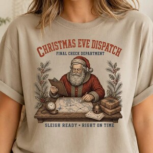 May include: A beige t-shirt featuring a graphic of Santa Claus at a desk with a map, a clock, and gifts. The text reads: CHRISTMAS EVE DISPATCH, FINAL CHECK DEPARTMENT, SLEIGH READY, RIGHT ON TIME. The design includes pine branches and holly.