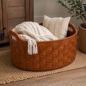 Pode incluir: Um cesto redondo de couro entrançado em tom castanho com duas pegas laterais integradas. Dentro do cesto encontra-se uma manta de malha grossa em tom creme com franjas e uma pequena almofada decorativa retangular com riscas beges e brancas.