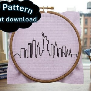 May include: A wooden embroidery hoop containing light purple Aida fabric with a black cross-stitched outline of the New York City skyline, including the Statue of Liberty. The image includes the text PDF Pattern Instant download.