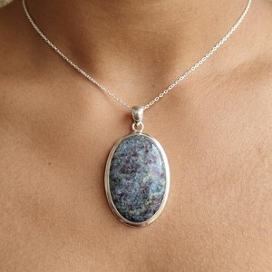 May include: A silver necklace featuring an oval-shaped pendant with a mottled grey and deep red stone. The pendant is set in a smooth silver bezel and hangs from a delicate silver chain that rests against the collarbone area of the neck.