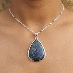 May include: A teardrop-shaped pendant necklace featuring a dark blue and grey stone with speckled inclusions and a vibrant magenta-pink centre. The stone is set in a polished silver-toned metal bezel and hangs from a delicate silver-toned chain.