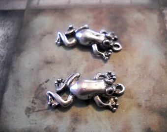 Pode incluir: Dois pingentes de sapo prateados com um laço para prender em joias ou outras artesanato.