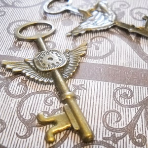 May include: Two ornate antique-style keys with wings. One key has a clock face on the bow. Both keys are made of gold-toned metal.