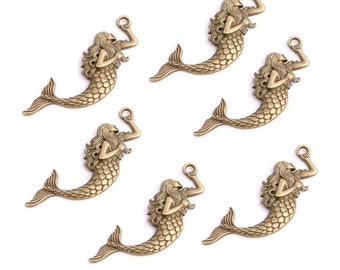 May include: Six antique gold-toned mermaid charms with a raised design. Each charm features a mermaid with flowing hair and a tail, holding a seashell in her hand.