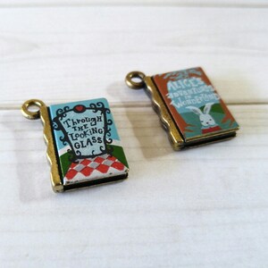 May include: Two antique gold-toned book charms with Alice in Wonderland themes. One charm features the text "Alice's Adventures in Wonderland" and a white rabbit illustration. The other charm features the text "Through the Looking Glass" and a checkered pattern.
