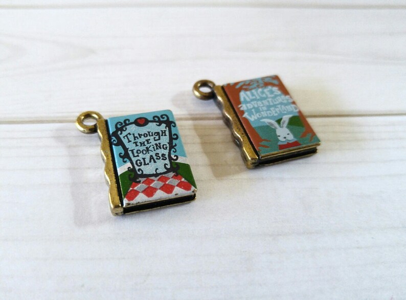 May include: Two antique gold-toned book charms with Alice in Wonderland themes. One charm features the text "Alice's Adventures in Wonderland" and a white rabbit illustration. The other charm features the text "Through the Looking Glass" and a checkered pattern.