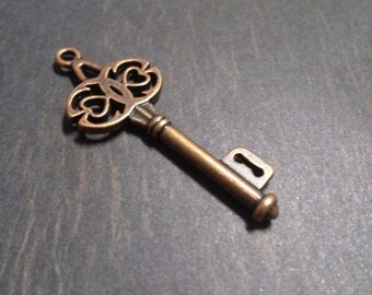 May include: Bronze-colored key with an ornate, openwork design on the head. The key has a long, cylindrical stem and a keyhole-shaped opening. The key is likely a decorative item or charm.