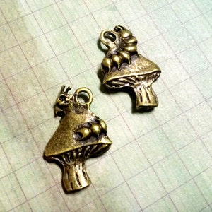 May include: Two antique gold-toned mushroom charms with a small bug on top. The charms have a detailed design and are perfect for jewelry making or crafting.