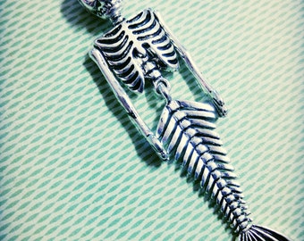 May include: Silver skeleton mermaid charm with a detailed ribcage and a fishtail. The charm is on a green and white patterned background.
