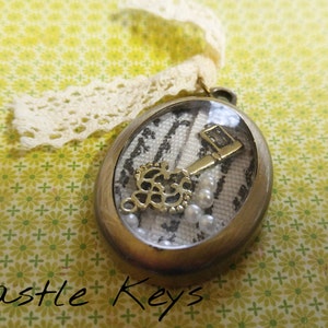 May include: A gold key charm encased in a clear resin pendant with a white and black striped fabric background. The pendant is attached to a white lace ribbon. The text "Castle Keys" is visible below the pendant.