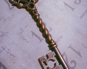 May include: A large, ornate, antique-style key made of copper metal. The key has a decorative, intricate head and a twisted, ornate shaft. The key is lying on a white surface with numbers printed on it.
