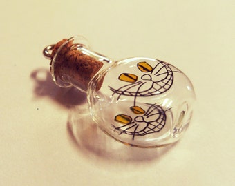 May include: A small glass bottle with a cork stopper and a painted image of a Cheshire Cat with a wide grin and yellow eyes. The bottle is clear and has a textured surface.