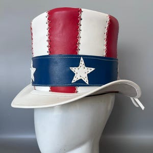 May include: A top hat featuring vertical red and white leather panels stitched together with white thread. A wide blue leather band encircles the base, decorated with white leather stars. The hat has a curved white brim and a white leather tie.