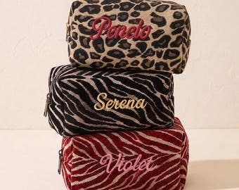 May include: Three rectangular zippered pouches stacked vertically. The top pouch features a tan and black leopard print with the name Penelo embroidered in red. The middle pouch has a black and white zebra print with the name Serena in gold. The bottom pouch is a red and white zebra print with the name Violet in pink.