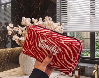 May include: A rectangular zippered pouch featuring a red and beige zebra stripe pattern. The name Emily is embroidered in white cursive script on the front center of the pouch. The bag has a structured shape with a black zipper closure along the top edge.