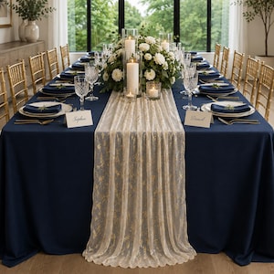 May include: A long, sheer, cream-colored table runner with a delicate gold floral pattern draped over a solid navy blue tablecloth. The table is set with white plates, navy blue napkins, crystal glassware, and place cards labeled Sophia and Daniel.