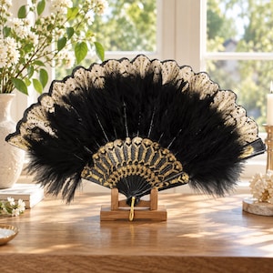 May include: A handheld folding fan featuring black feathers and a black frame with intricate gold-colored detailing. The top edge of the fan is finished with a decorative gold-colored lace trim. The fan is displayed open on a small wooden stand.