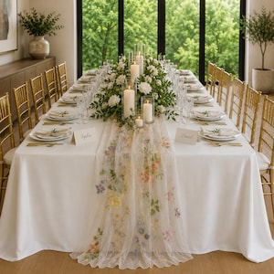 May include: A long rectangular dining table covered with a white tablecloth and a sheer floral embroidered table runner. The table is set with white plates, gold cutlery, clear glassware, place cards, and a centerpiece of white flowers and candles.