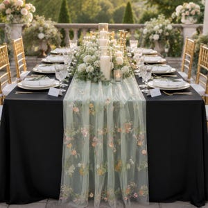 May include: A black tablecloth draped over a rectangular table, featuring a sheer sage green tulle table runner embroidered with delicate floral patterns. The table is set with white plates, clear glassware, gold cutlery, and white place cards.