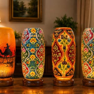 May include: Four cylindrical table lamps with illuminated bodies on circular wooden bases. The first lamp features a black silhouette of a camel rider in a desert landscape. The other three lamps display colorful, repeating floral patterns.