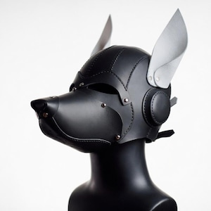 Peut inclure: Un masque de tête en forme d'animal noir doté d'un museau pointu, d'oreilles droites proéminentes et de panneaux latéraux circulaires. Le masque est construit avec des coutures visibles et des rivets métalliques, couvrant la tête.