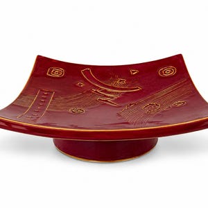 May include: A square, deep red ceramic dish with curved, upturned edges and a circular pedestal base. The surface features etched, gold-toned linear patterns, including swirls, dashes, and textured lines, with a thin gold rim along the edges.