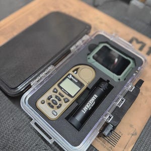 May include: A tan Kestrel ballistics meter, a black Athlon Rangecraft cylinder, and a green device stored in a clear protective case with black foam inserts. The Kestrel device features a digital screen, navigation buttons, and the word Link.