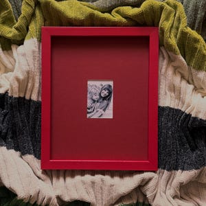 Pode incluir: Uma moldura retangular vermelha com um passe-partout vermelho escuro e uma pequena fotografia a preto e branco de duas pessoas centrada. A moldura tem um design de bordas planas e limpas e está posicionada verticalmente.