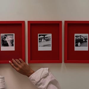 Può includere: Tre cornici rettangolari rosse montate su una parete, ognuna contenente una fotografia quadrata in bianco e nero. La foto a sinistra mostra un'auto, quella centrale una persona sorridente e quella a destra un'auto con un passeggero.