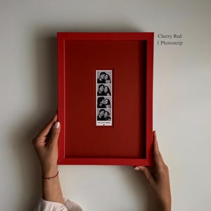 Puede incluir: Un marco de fotos rectangular de color rojo cereza que contiene una tira de fotos vertical en blanco y negro con cuatro imágenes de dos personas. El marco incluye un paspartú rojo a juego. El texto a la derecha dice Cherry Red 1 Photostrip.