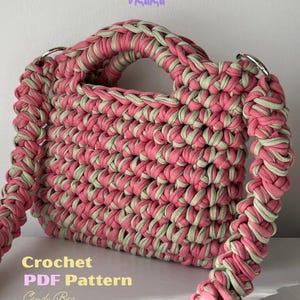 Può includere: Una borsa fatta all'uncinetto a mano con filo di tessuto spesso intrecciato rosa e verde chiaro. La borsa presenta un corpo rettangolare, manici integrati e una tracolla intrecciata fissata con anelli in metallo argentato. Testo: Crochet PDF Pattern Candy Bag.