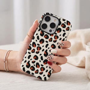 Op de afbeelding: Een smartphonehoesje met een crèmekleurige achtergrond en een herhalend luipaardpatroon in zwart en gebrand oranje. Het hoesje heeft een rechthoekige uitsparing voor de cameralenzen en is ontworpen voor een telefoon met drie lenzen.