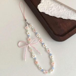 May include: A phone charm featuring a loop of white, pink, and light blue beads arranged in a floral pattern. The accessory includes a soft pink fabric ribbon tied into a bow and a thin pink cord attachment for securing to a mobile device.
