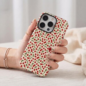 Op de afbeelding: Een smartphonehoesje met een herhalend patroon van kleine rode kersen met groene steeltjes op een crèmekleurige achtergrond. Het hoesje heeft een zwarte uitsparing voor de camera en bedekt de achterkant en zijkanten van het mobiele apparaat.