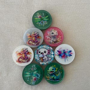 May include: A collection of eight circular resin cabochons featuring colorful illustrations of dragons, axolotls, and dragonflies. The designs include a green dragonfly, a purple dragon, a pink dragon, a white dragon, a blue axolotl with ice cream, a blue dragonfly, a green floral dragonfly, and a green dragon.