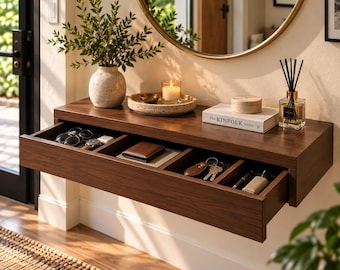 May include: A dark wood floating wall console with a single wide drawer divided into four compartments. The drawer contains sunglasses, a watch, a wallet, keys, and a pen. The top surface holds a ceramic vase, a candle, and a book titled The Kinfolk.